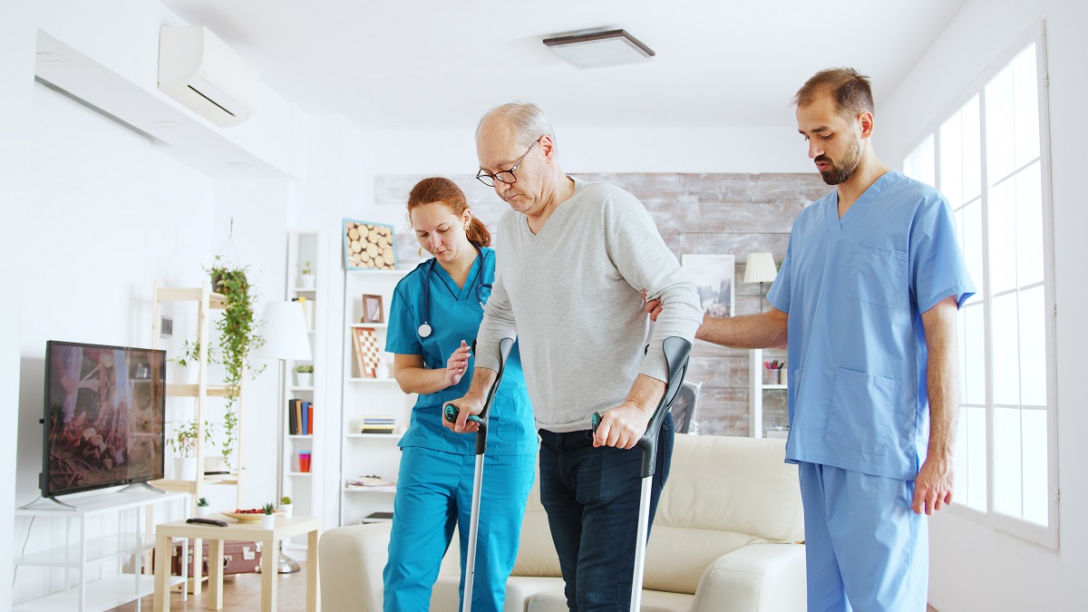 Team of nurses or social workers helping an old disabled man to walk with his crutches out of the nursing home room